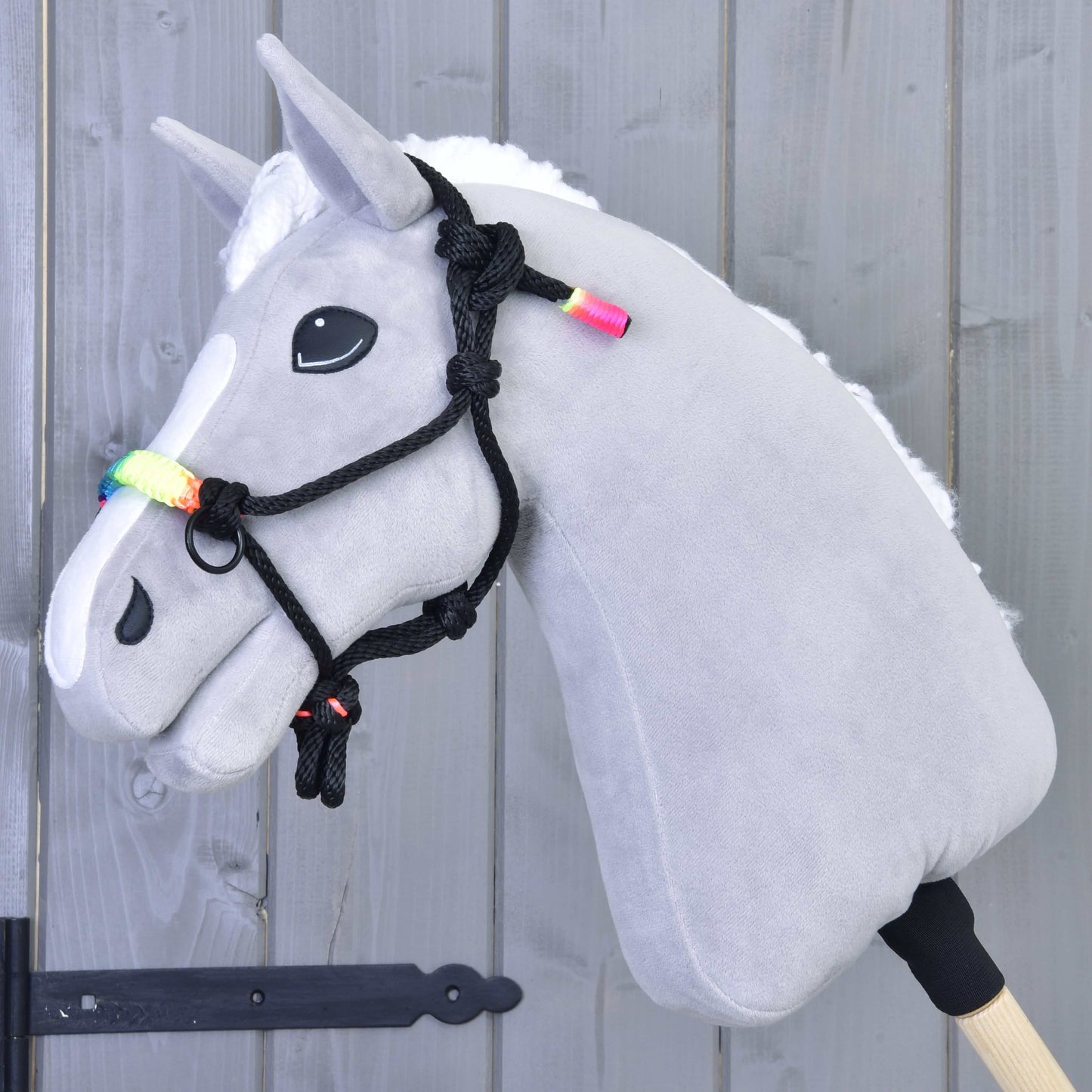 Parelka Dúha od Hobby Horse LarDen na sivom Hobby Horse s bielou hrivou, dúhovo farebnými korálkami a čiernou povrazovou ohlávkou na drevenej tyči, pred svetlosivou drevenou stenou.