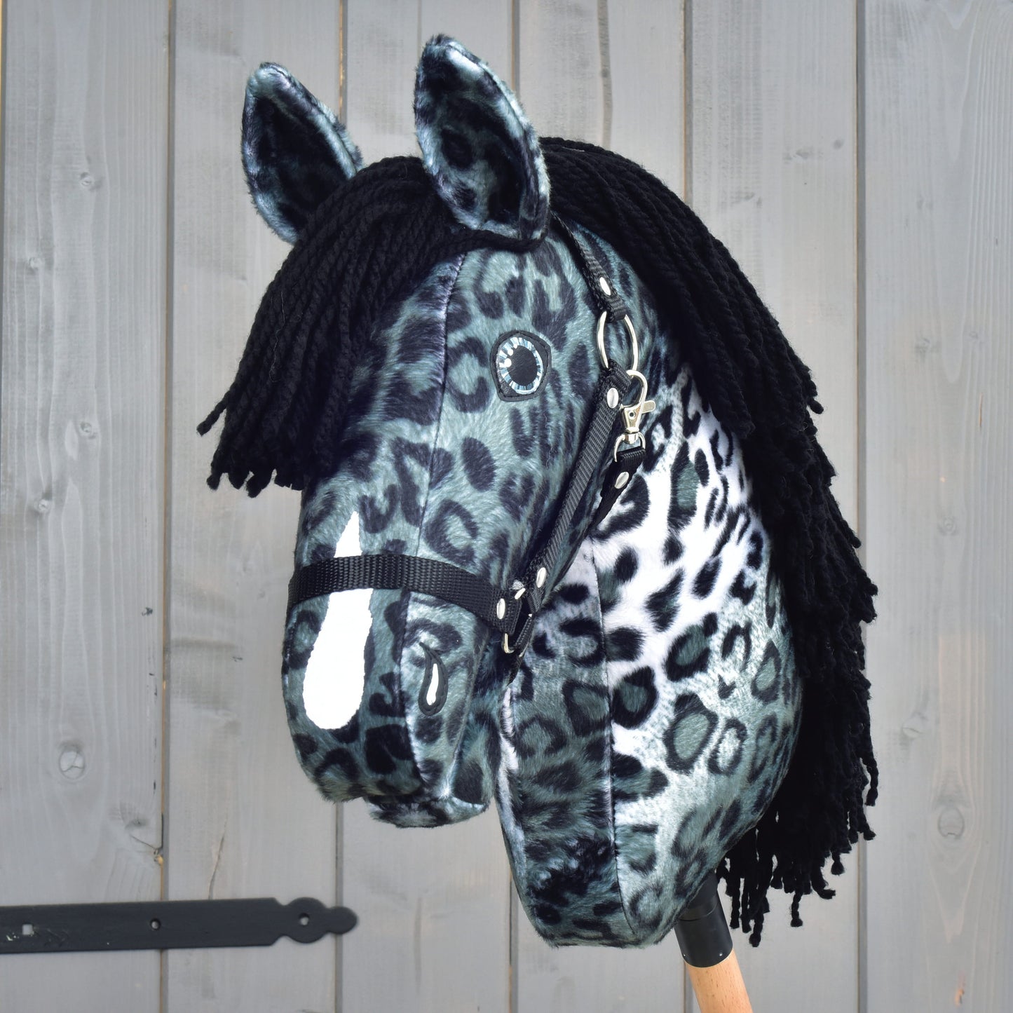 Hobby Horse Picasso s čiernou ohlávkou od Hobby Horse LarDen, plyšový Hobby Horse navrhnutý tak, aby pripomínal žrebca appaloosy, s výrazným čiernobielym leopardím vzorom, ktorý odkazuje na vzhľad škvrnitej srsti. Má čiernu priadzovú hrivu, čiernu ohlávku a je upevnený na drevenej tyči, na pozadí drevených panelov.