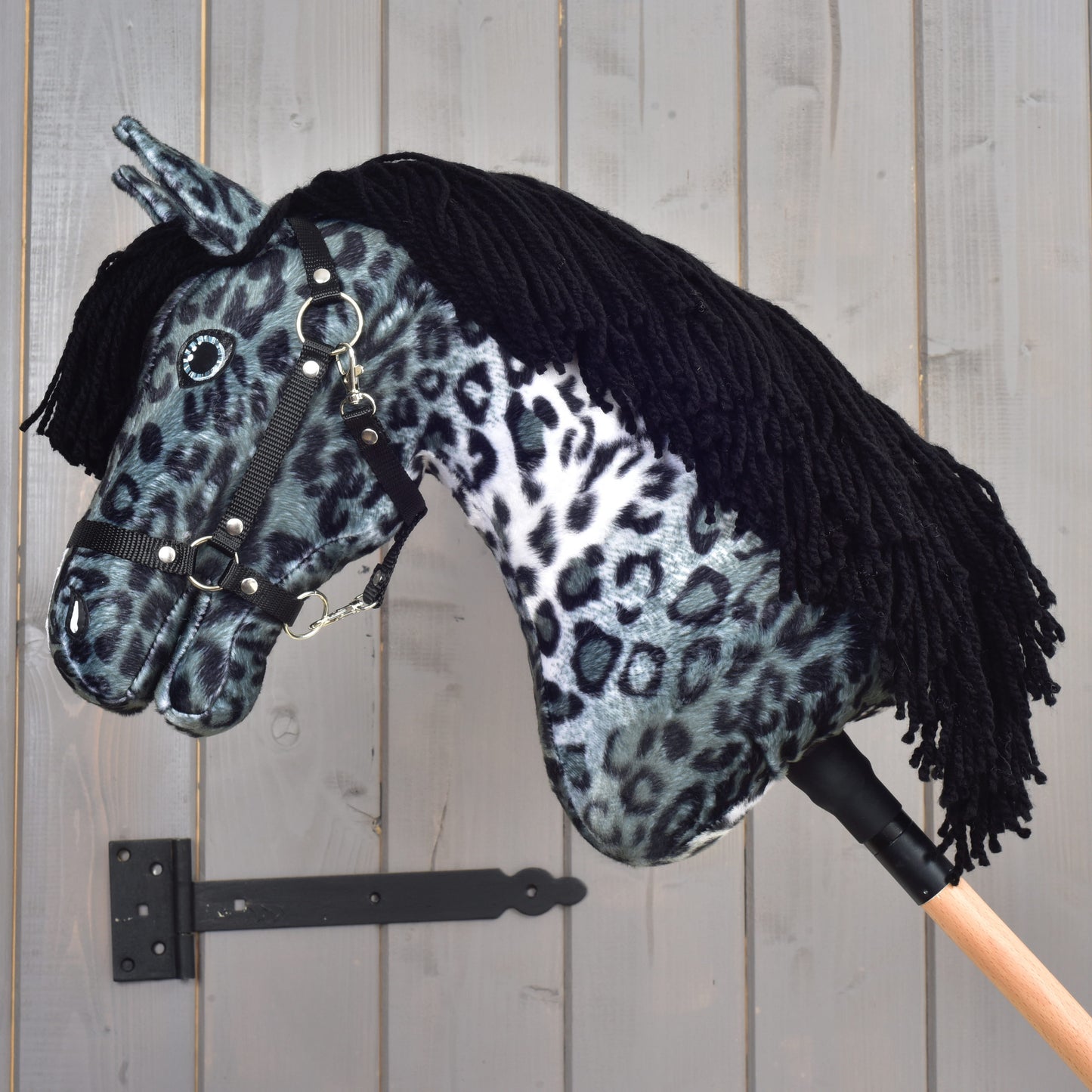 Hobby Horse Picasso s čiernou ohlávkou od Hobby Horse LarDen s plyšovou hlavou so sivočiernou škvrnitou srsťou inšpirovanou appaloosou, čiernou ohlávkou a čiernou priadzovou hrivou, upevnený na drevenej tyči a zobrazený na pozadí sivej drevenej steny.
