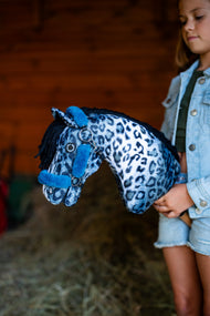 Dievča v džínsovej bunde drží Hobby Horse Picasso s čiernou ohlávkou od LarDen pri červenej drevenej stene stodoly, modrobiela plyšová hlava s leopardím vzorom a čierna priadzová hriva, s ohlávkou Teddy Petrol s kožušinou
