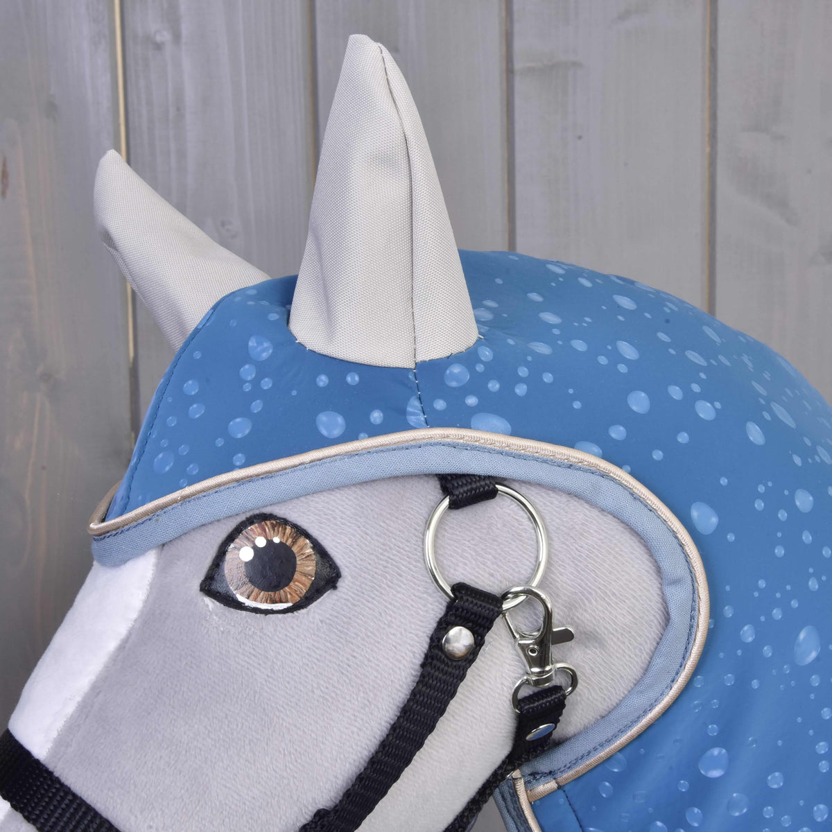 Detail hlavy Hobby Horse LarDen Pláštenka Dolly (softshell) s modrou pláštenkou so vzorom kvapiek vody a čiernou ohlávkou, na pozadí svetlých drevených panelov – ideálna na hru za každého počasia.