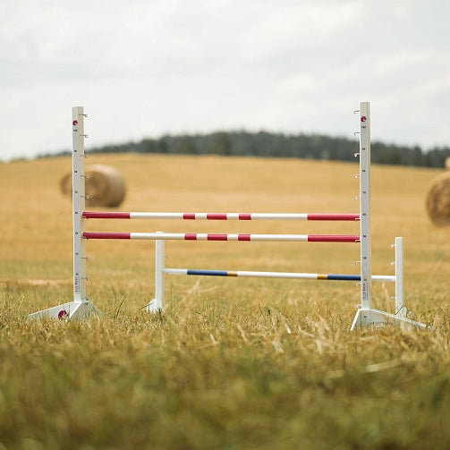 Biele skokové tyče s červenými pruhmi postavené na trávnatom poli, ideálne pre Hobby Horsing, s balíkmi sena a stromami viditeľnými v pozadí pod zatiahnutou oblohou.
