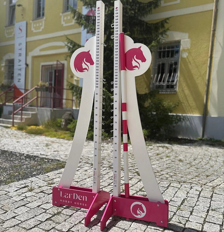 Dva biele a ružové nastaviteľné stojany na skoky pre Hobby Horsing sú postavené vonku na dláždenej ceste pred žltou budovou so schodmi a ceduľou v pozadí.