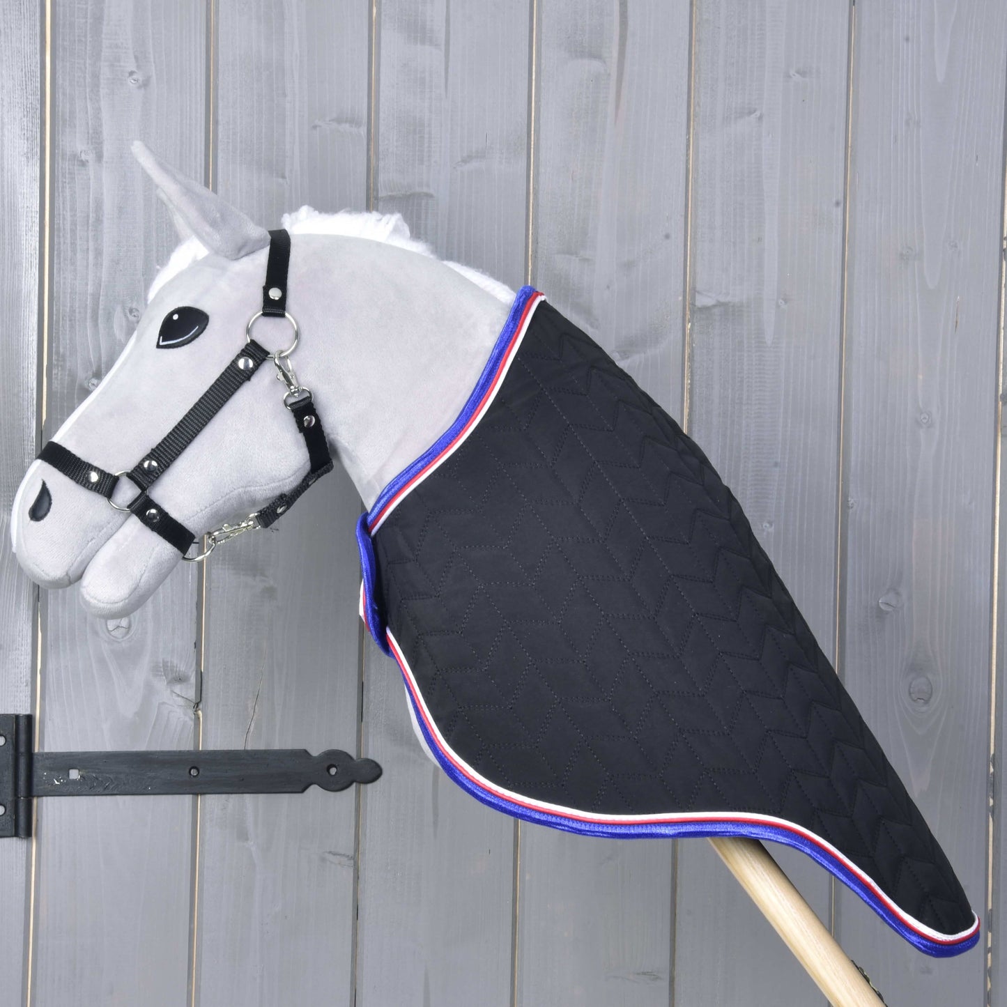 Šedá hračka Hobby Horse LarDen s převozní dekou Luxury — černou prošívanou dekou s červeno-bílo-modrým lemováním — stojí na dřevěné tyči na rustikálním dřevěném pozadí.