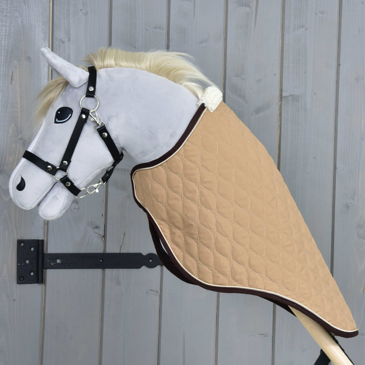 Plyšový Hobby Horse so svetlosivou hlavou, bielou hrivou a čiernou uzdečkou má na sebe prepravnú deku Paris od Hobby Horse LarDen — ľahkú odolnú pieskovú prešívanú deku — je na drevenej tyči a na pozadí sivej drevenej steny.