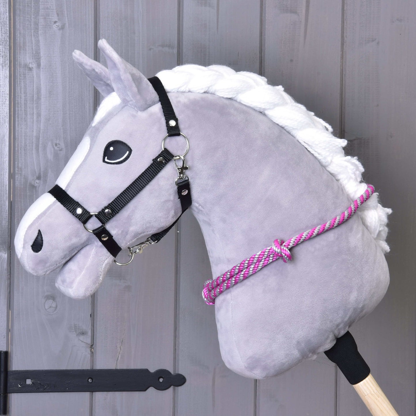 Nákrčník s ružovým prúžkom od Hobby Horse LarDen na sivej plyšovej konskej hlave s bielou hrivou, čiernou uzdečkou a ružovo-bielym nákrčníkom na drevenej tyči, ideálny pre nácvik jazdy bez uzdečky pred sivou drevenou stenou.