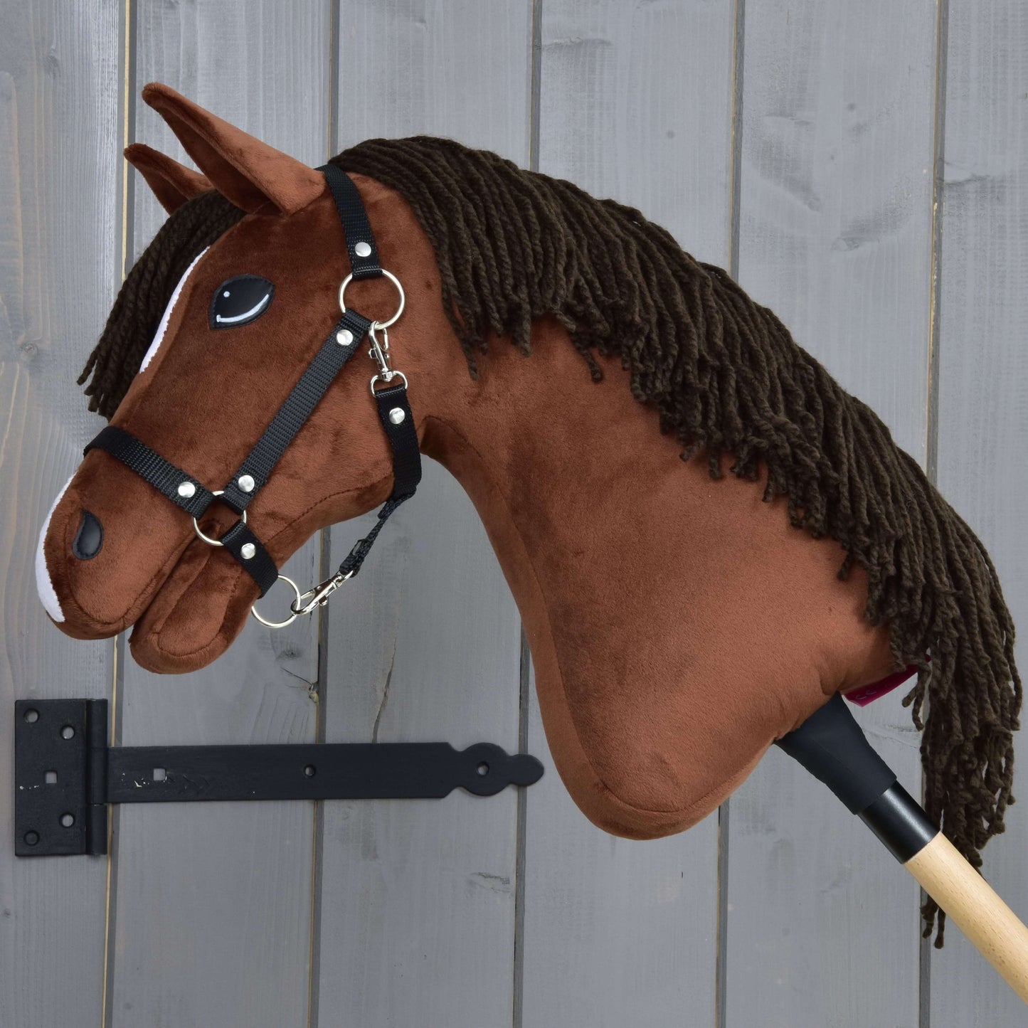 Hobby Horse Rocky s černou ohlávkou od Hobby Horse LarDen, hnědý plyšový kůň na tyči, je zobrazen vedle šedé dřevěné stěny.