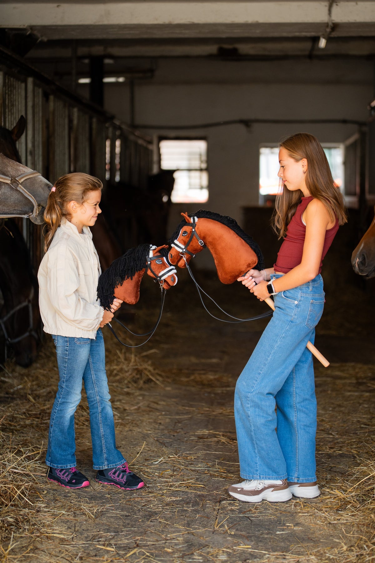 Dve dievčatá stoja oproti sebe v uličke stajne a každé drží Hobby Horse Rocky od Hobby Horse LarDen s gaštanovohnedou plyšovou hlavou s bielou lysinou a čiernou hrivou z priadze, s čiernou kompletnou uzdečkou s kožušinou a opratami