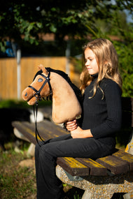 Dievča v čiernom svetri sedí na lavičke v parku vo večernom slnku a drží Hobby Horse Samantha with Black Halter od LarDen – pieskovú plyšovú hlavu s čiernou priadzovou hrivou – s čiernou uzdečkou s remienkovými oťažami
