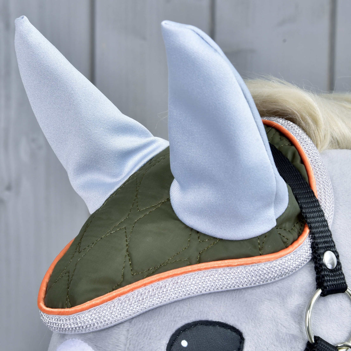 Set Moos od Hobby Horse LarDen, detail hlavy plyšového koňa s olivovo zelenou a oranžovou čabrakou, dvoma svetlosivými špicatými látkovými ušami a roztomilou konskou dekoráciou na svetlosivom drevenom pozadí.