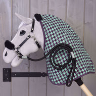 Set Harmony Green od Hobby Horse LarDen – plyšový Hobby Horse s čiernou ohlávkou, zeleno-fialovou stajňovou dekou so vzorom kohútej stopy a stočeným čiernym vodidlom – je zavesený na drevenom nástennom držiaku pred sivou drevenou stenou.