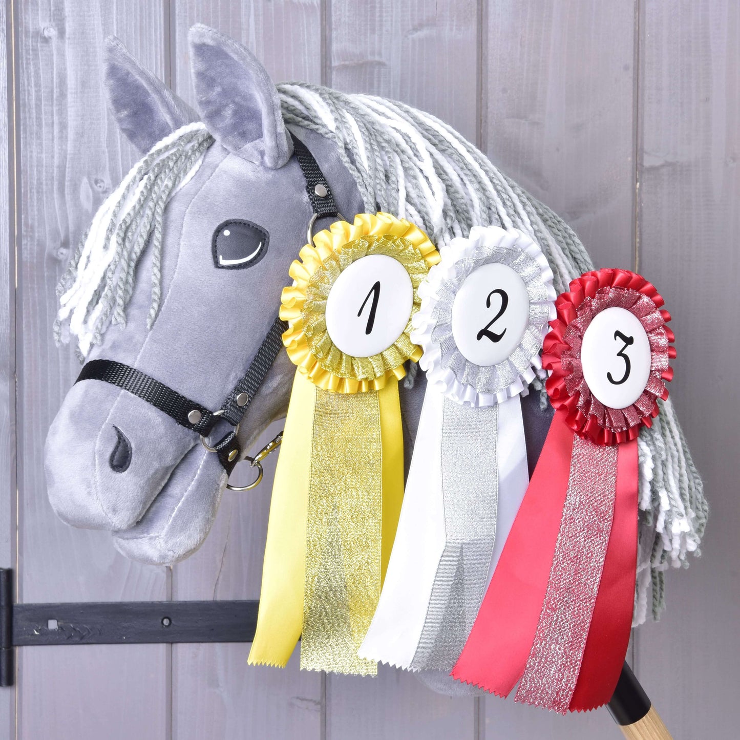 Sivý plyšový Hobby Horse s bielou priadzovou hrivou má na krku zavesený Hobby Horse LarDen Set Hobby Horse Kokarda NEW – žltú, bielu a červenú kokardu pre prvé, druhé a tretie miesto – na drevenom pozadí.