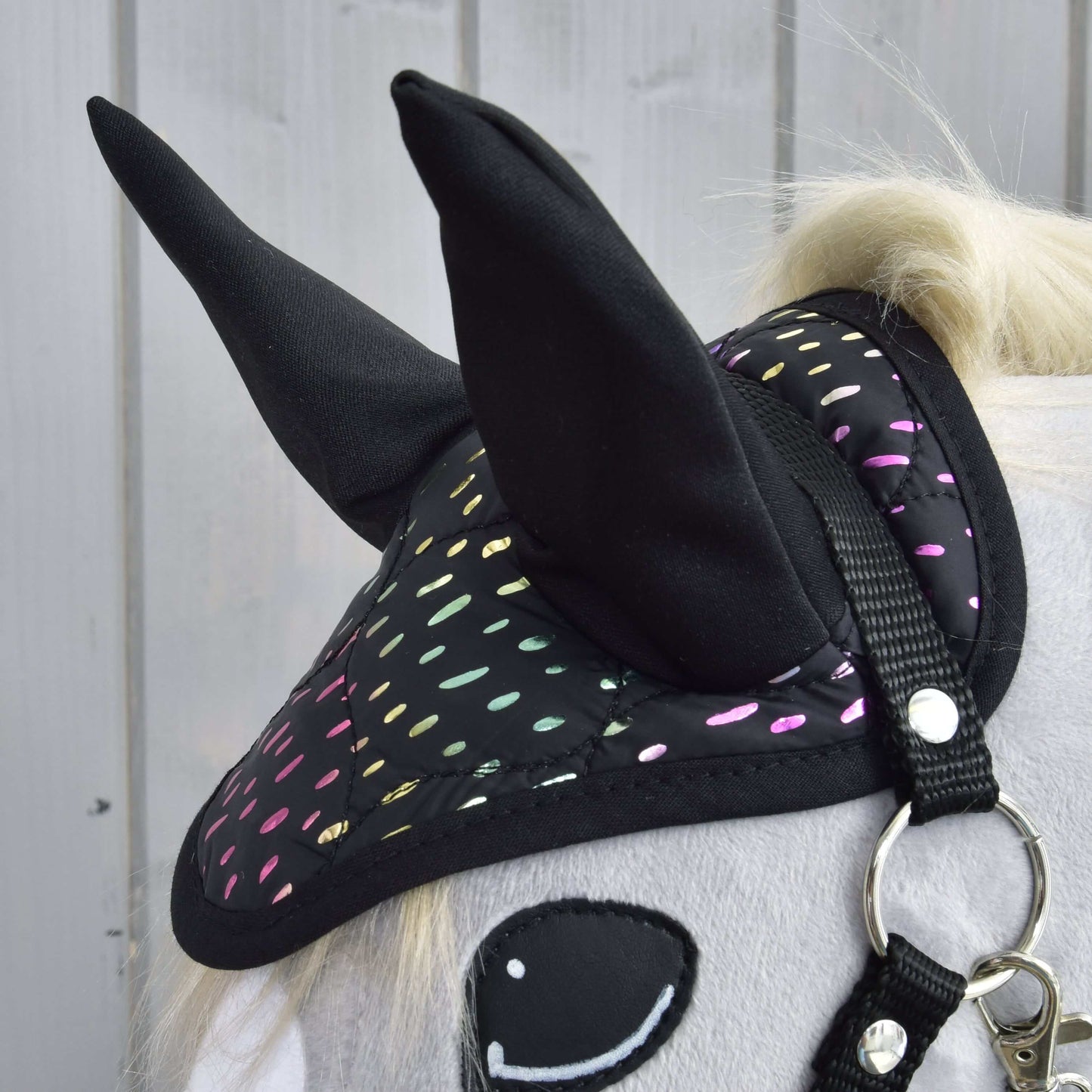 Detail Hobby Horse LarDen „Set Dreamy“ na Hobby Horse s bielym telom, čiernou čabrakou s farebnými reflexnými bodkami, usmievavým plsteným okom a uzdečkou, pred svetlým dreveným obkladom.