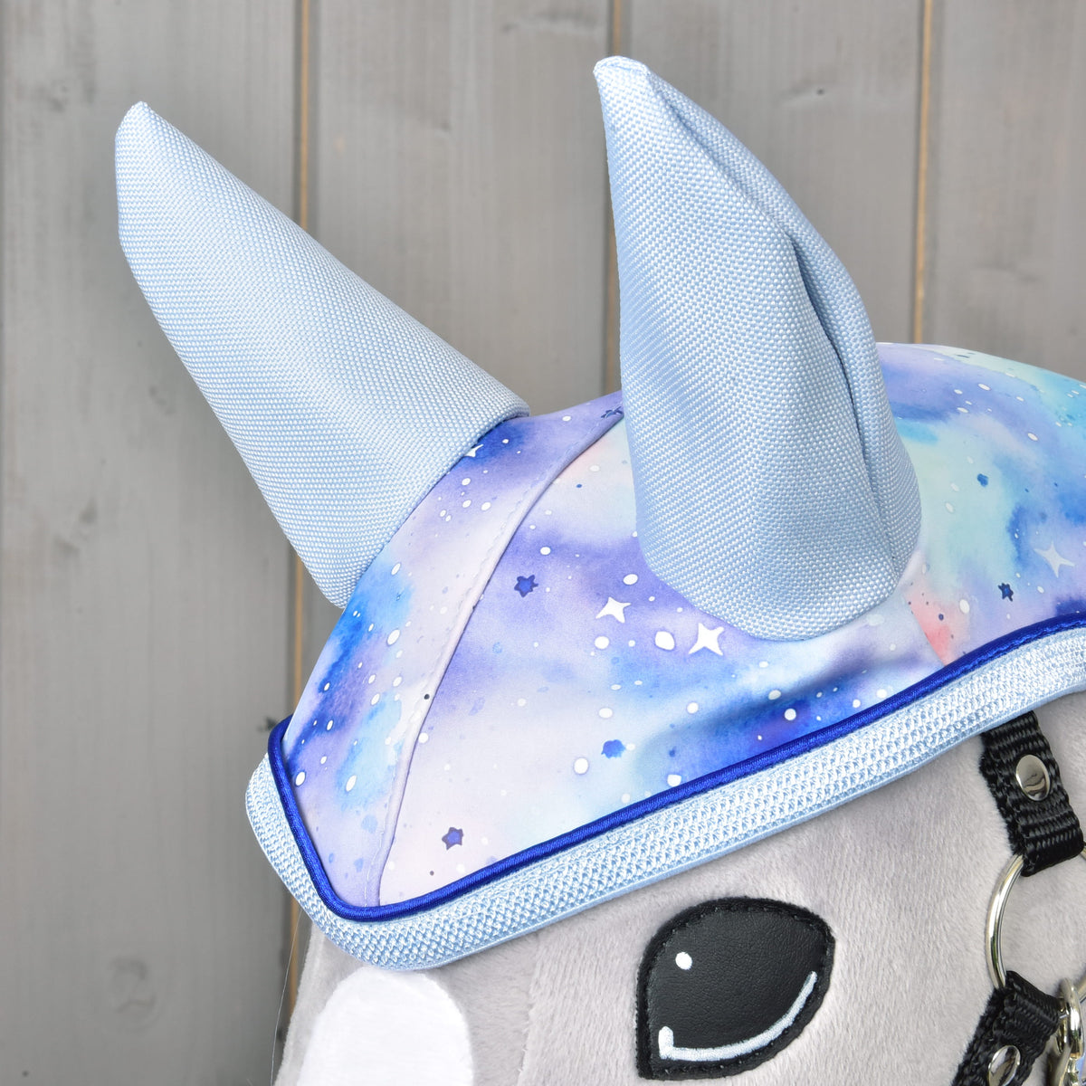 Detail Hobby Horse LarDen Pláštenka Space: plyšové zvieratko má na sebe čiapočku s vysokými svetlomodrými rohmi a modrofialovým galaktickým vzorom na drevenom pozadí – ideálne zobrazenie tohto doplnku v podobe pláštenky do každého počasia.