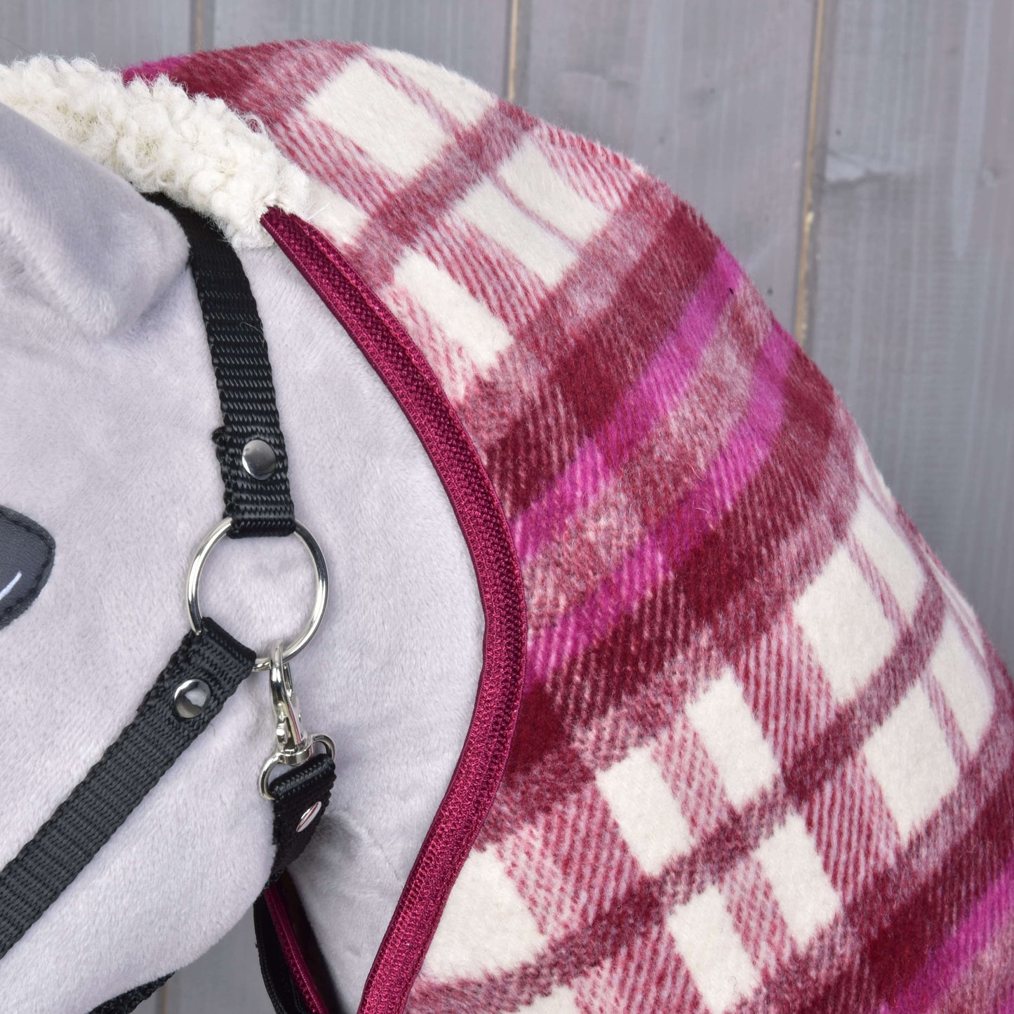 Deka stajňová LarDen od Hobby Horse LarDen na sivom koni pred drevenými doskami, v červeno-ružovo-bielej kocke, s bielou fleecovou podšívkou a ohlávkou.