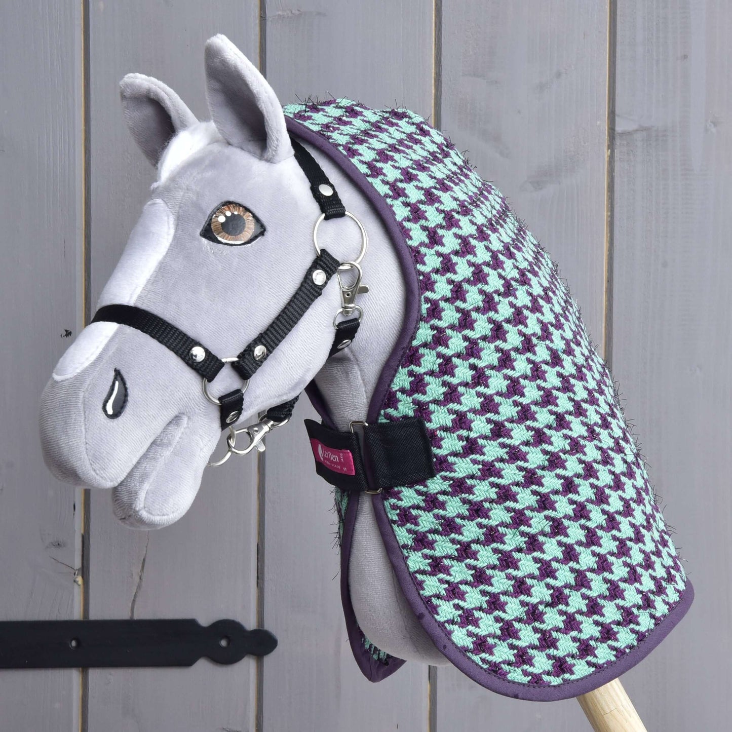 Ručne vyrobený sivý Hobby Horse s čiernou uzdečkou, oblečený v deke Hobby Horse LarDen „Deka stajňová Green“, vystavený pred dreveným pozadím.