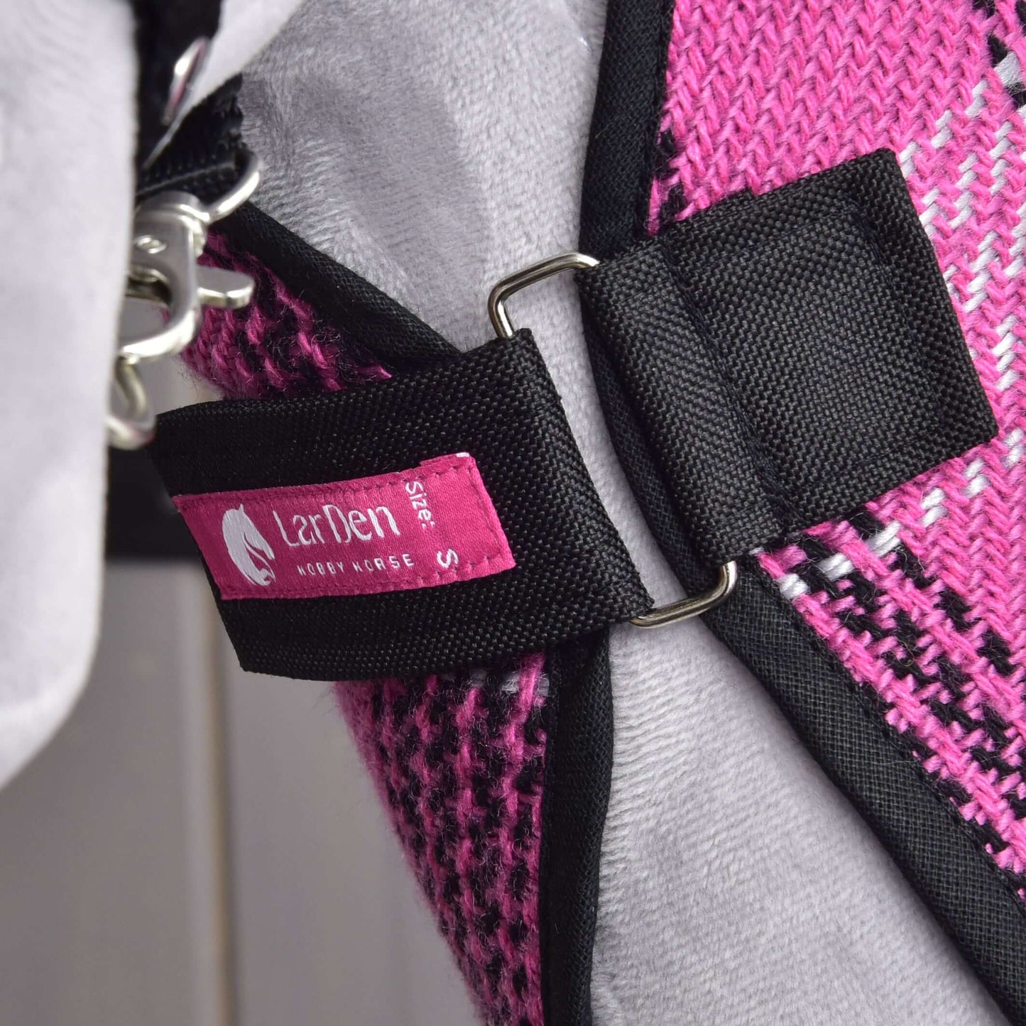 Detail ružovo-čierneho látkového postroja s kovovým krúžkom a štítkom Hobby Horse LarDen Stable Blanket Pink Size S, pripevneného k sivému materiálu, možno ako súčasť tréningovej výbavy pre kone alebo deky pre Hobby Horse, nosenej domácim maznáčikom.