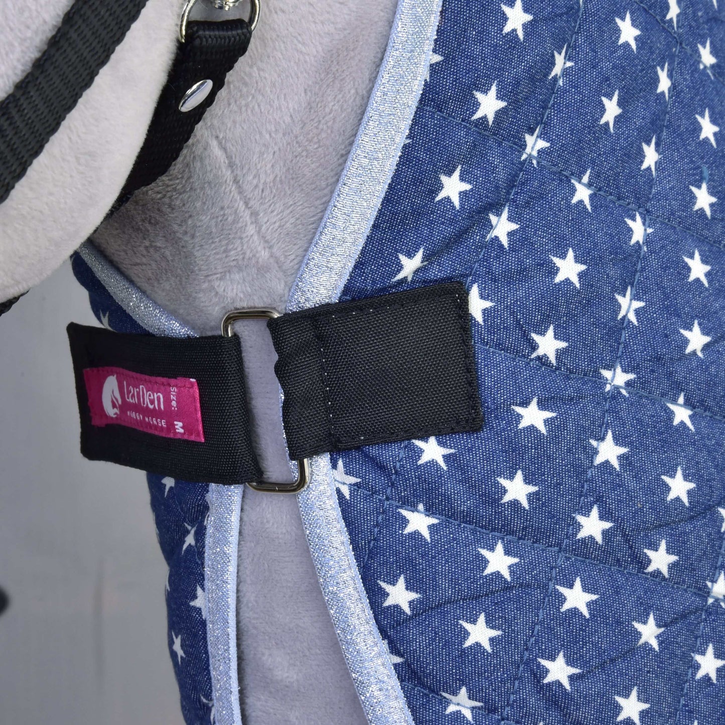 Detail modrej prešívanej deky Deka stajňová Star od Hobby Horse LarDen s bielymi hviezdami, čiernym pásikom so striebornou prackou a ružovým štítkom Izefan, nosenej sivým zvieraťom ako súčasť jeho postroja alebo vesty.