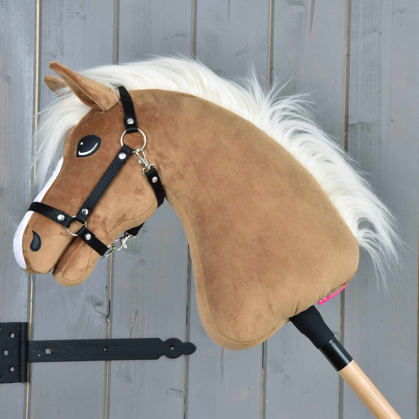 Hobby Horse Star s čiernou ohlávkou od Hobby Horse LarDen, s hnedou plyšovou hlavou, karamelovou srsťou, bielou hrivou, čiernou ohlávkou a bielymi znakmi na nose, na drevenej tyči pred stenou zo sivých drevených dosiek.