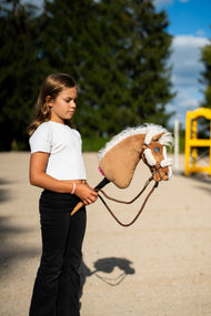 Dievča v bielom tričku vedie Hobby Horse Star with black halter od LarDen cez pieskové kolbisko, za ním je žlté krídlo prekážky – palominová plyšová hlava s vlajúcou bielou priadzovou hrivou – s hnedou ohlávkou s úplným kožušinovým lemom a oťažami Vivaldi