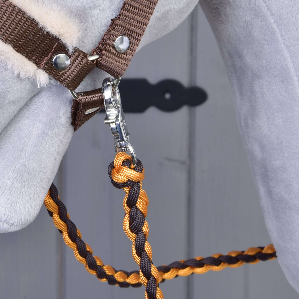 Detailný záber na konskú hlavu so Set Perfect Combination Brown od Hobby Horse LarDen, s ohlávkou s kožušinovým polstrovaním a oranžovo-hnedým pleteným vodidlom so striebornou karabínou. V pozadí sú rozmazané sivé dvere s čiernym štítkom kľúčovej dierky – ideálny pre fanúšikov Hobby Horse.
