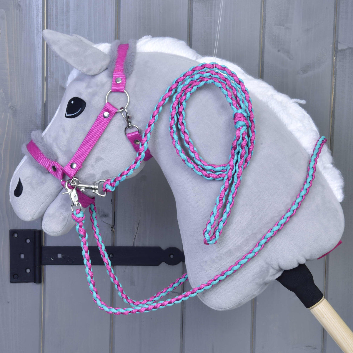 Set Perfect Combination Pink od Hobby Horse LarDen, sivý plyšový Hobby Horse s ružovou a tyrkysovou ohlávkou s kožušinovým polstrovaním a oprátami, visí na nástennom háčiku pred sivým dreveným pozadím.