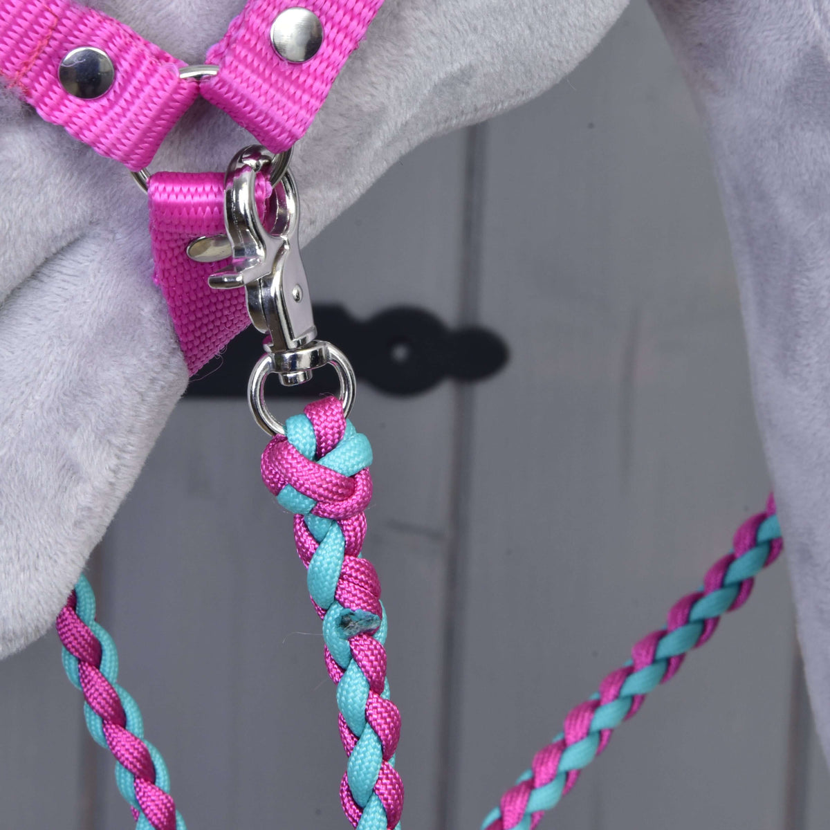 Detailný záber na Set Perfect Combination Pink od Hobby Horse LarDen – ružový postroj a ružovo-tyrkysové pletené vodidlo pripnuté ku krúžku na sivom povrchu – ideálny pre fanúšikov štýlových doplnkov. V pozadí vidno rozmazané svetlé drevo.