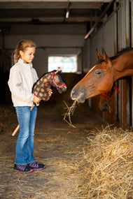 Dievča v krémovej bunde dvíha Hobby Horse Stormy s čiernou ohlávkou od LarDen k skutočnému hnedákovi žerúcemu seno v stajni, žíhaná hnedá plyšová hlava a čierna priadzová hriva, s ohlávkou Burgundy s kožušinou zo syntetickej kože