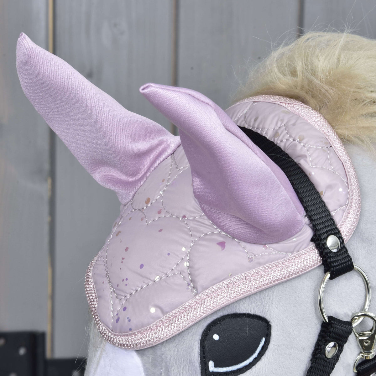 Detail Hobby Horse LarDen Set Sweetest Heart ukazuje jeho svetlosivú hlavu, lesklý prešívaný ružový jednorožčí roh, čabraku, čiernu uzdečku a drevené pozadie.