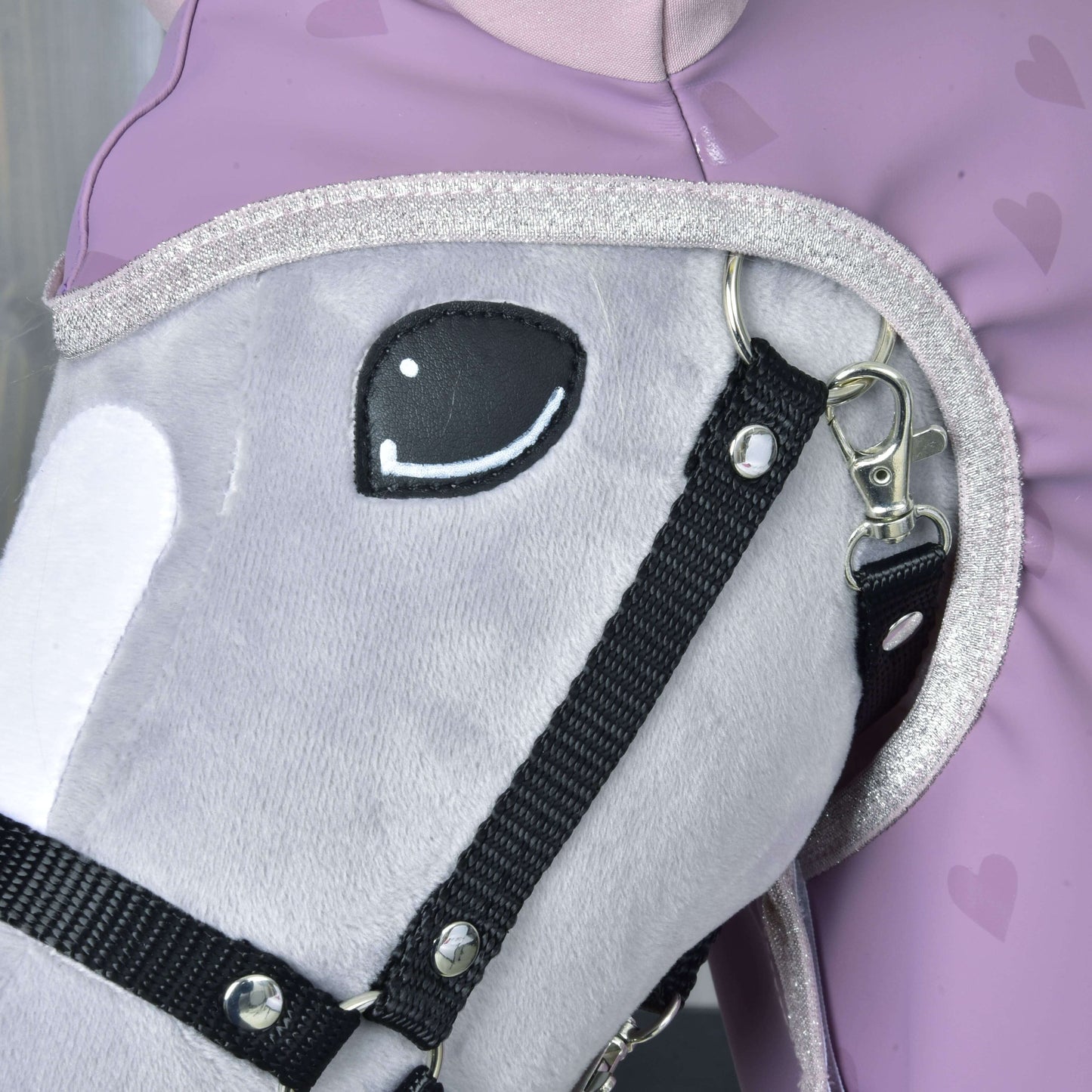Detail plyšového sivého Hobby Horse Hobby Horse LarDen s čiernou uzdečkou, s doplnkom Pláštenka Sweetheart, vedľa osoby v levanduľovom oblečení so srdiečkovým vzorom. Kôň má vyšívané usmievavé oko a trblietavý lem na krku.