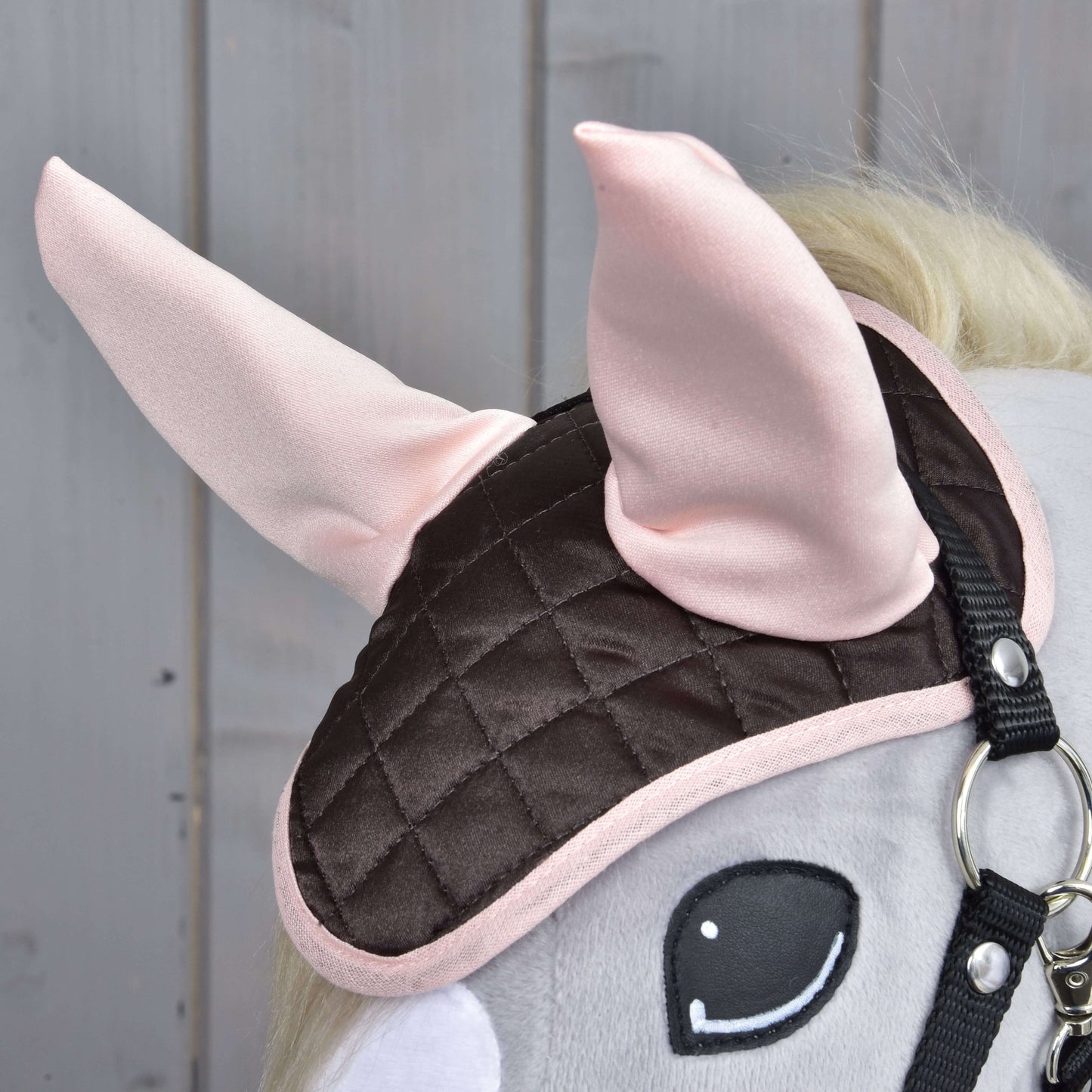 Detail Hobby Horse LarDen s čabrakou Chocolate, prešívanou hnedo-ružovou čabrakou so svetloružovými látkovými ušami na ochranu proti hmyzu a s pripevnenou uzdečkou, na pozadí dreveného obloženia.