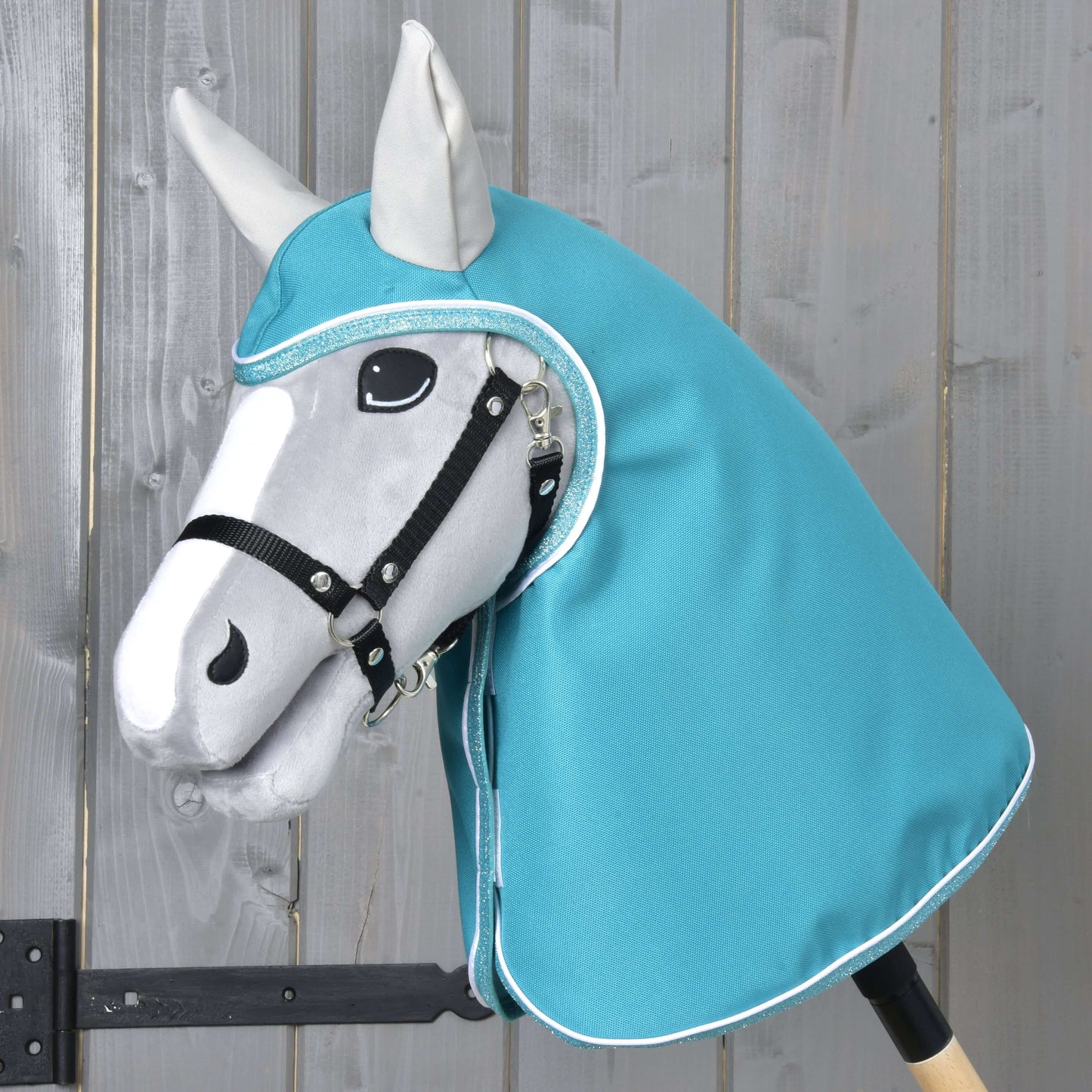 Pláštěnka Turquoise od Hobby Horse LarDen na šedém Hobby Horse s černou uzdečkou a tyrkysovou kapucí ve stylu pláštěnky, s realisticky tvarovanou hlavou s černými a bílými detaily, na světlém dřevěném pozadí.