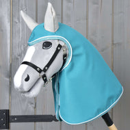 Pláštenka Turquoise od Hobby Horse LarDen na sivom Hobby Horse s čiernou uzdečkou a tyrkysovou kapucňou v štýle pláštenky, s realisticky tvarovanou hlavou s čiernymi a bielymi detailmi, na svetlom drevenom pozadí.