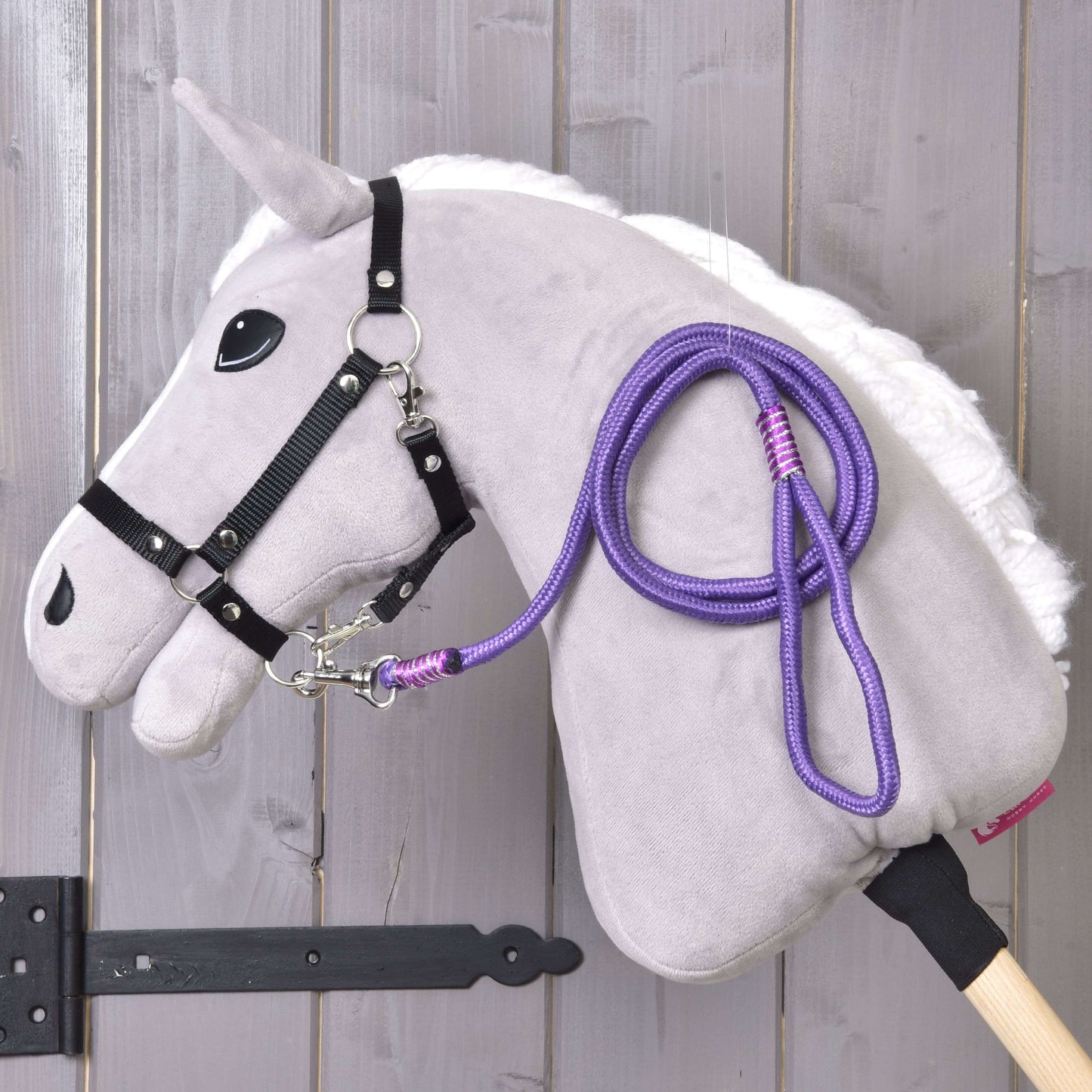 Hobby Horse - vodidlo Viola od Hobby Horse LarDen s plyšovým Hobby Horse, bielou hrivou, čiernou uzdečkou, fialovým povrazovým vodidlom a drevenou tyčou, zobrazené pred svetlou drevenou stenou.