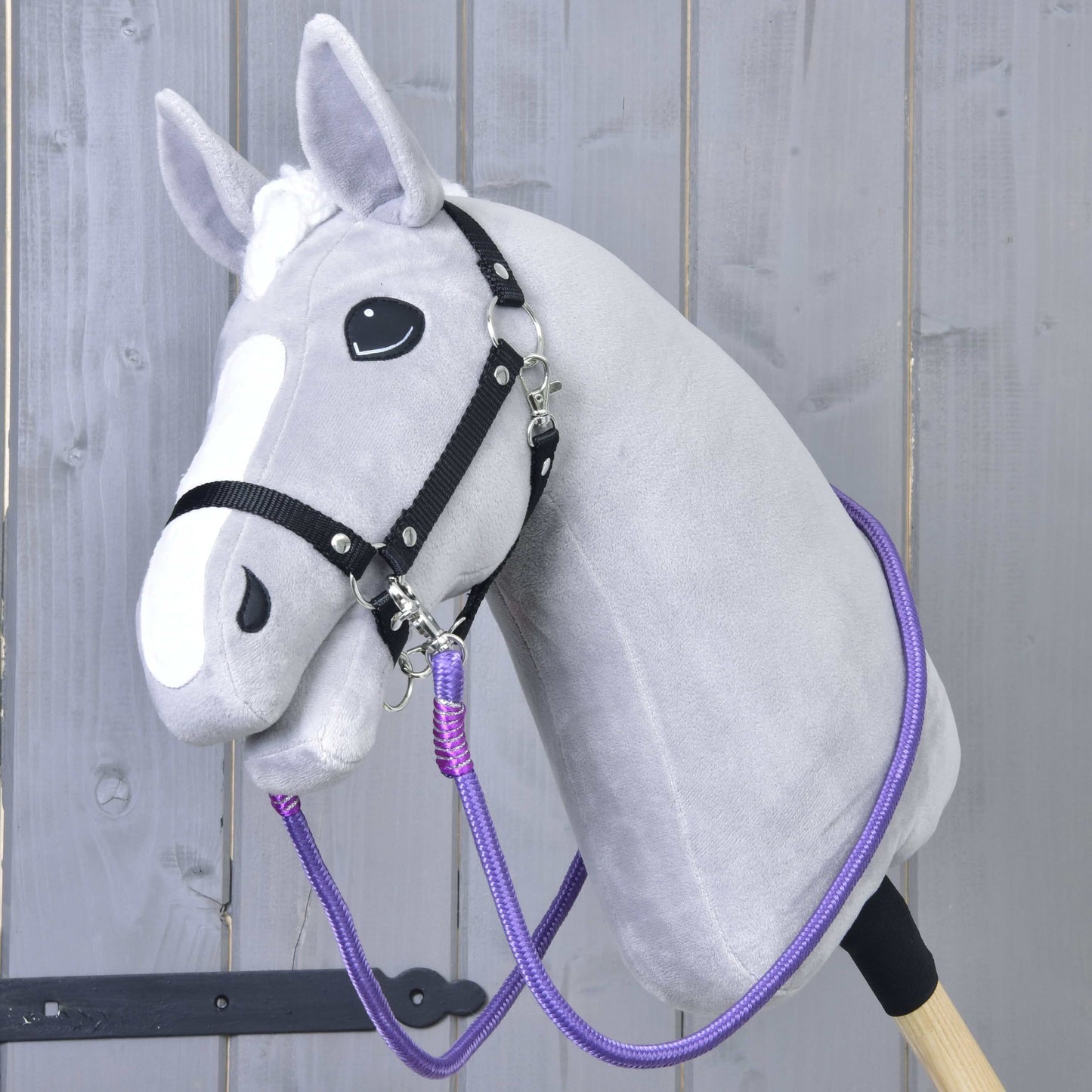 Hobby Horse LarDen „Opraty Viola“, sivý Hobby Horse s bielou lysinou, čiernou uzdečkou a výraznými ručne vyrábanými oprátami, s plyšovou hlavou na pevnej tyči, zobrazený pred svetlým dreveným plotom.
