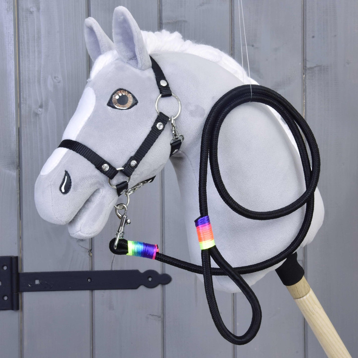 Hobby Horse - vodidlo Rainbow od Hobby Horse LarDen na sivom Hobby Horse s bielou hrivou, čiernou uzdečkou, opratami a vodidlom s dúhovou páskou okolo krku, vystavenom pred dreveným obložením.