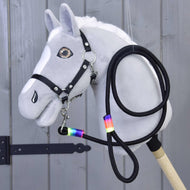 Hobby Horse - vodítko Rainbow od Hobby Horse LarDen na šedém Hobby Horse s bílou hřívou, černou uzdečkou, otěžemi a vodítkem s duhovou páskou kolem krku, vystaveném před dřevěným obložením.