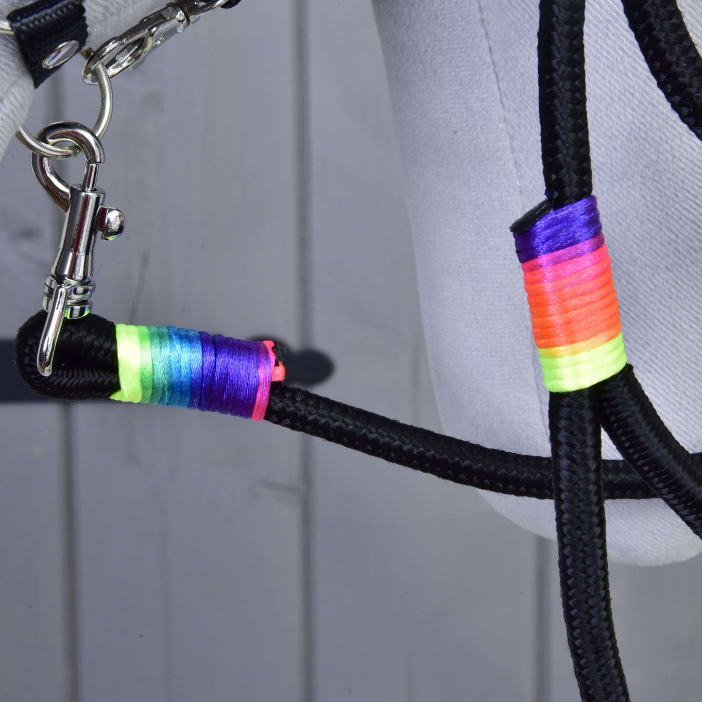 Detail Hobby Horse LarDen „Hobby Horse - vodidlo Rainbow“, čierneho pleteného vodidla s omotávkami z neónovo dúhovej nite a striebornou karabínou, pripevneného ku svetlosivej látke, na drevenom podklade.