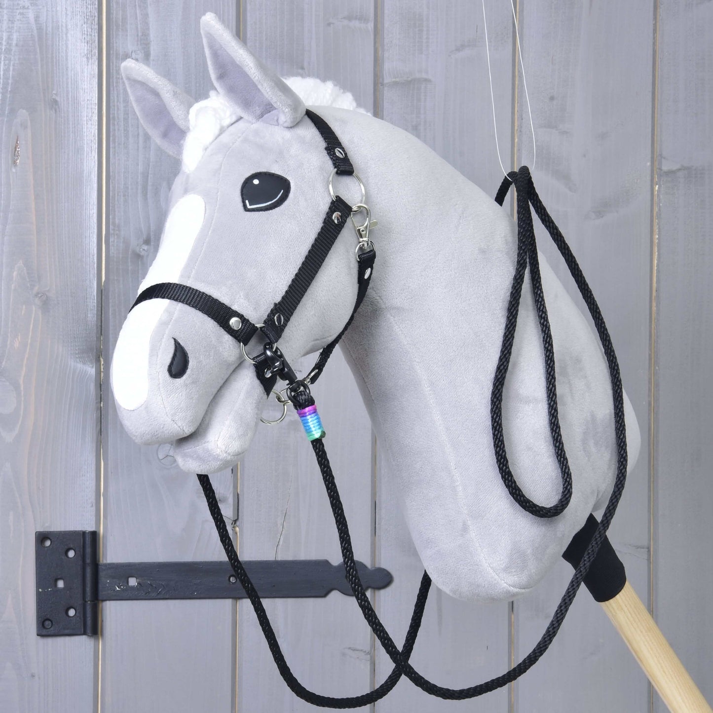 Hobby Horse LarDen "Westernové opraty Montana", sivý plyšový Hobby Horse s bielou hrivou a lysinou, čiernou uzdečkou s karabínami a westernovými oprátami, nasadený na pevnej drevenej tyči a zobrazený na sivej drevenej stene.