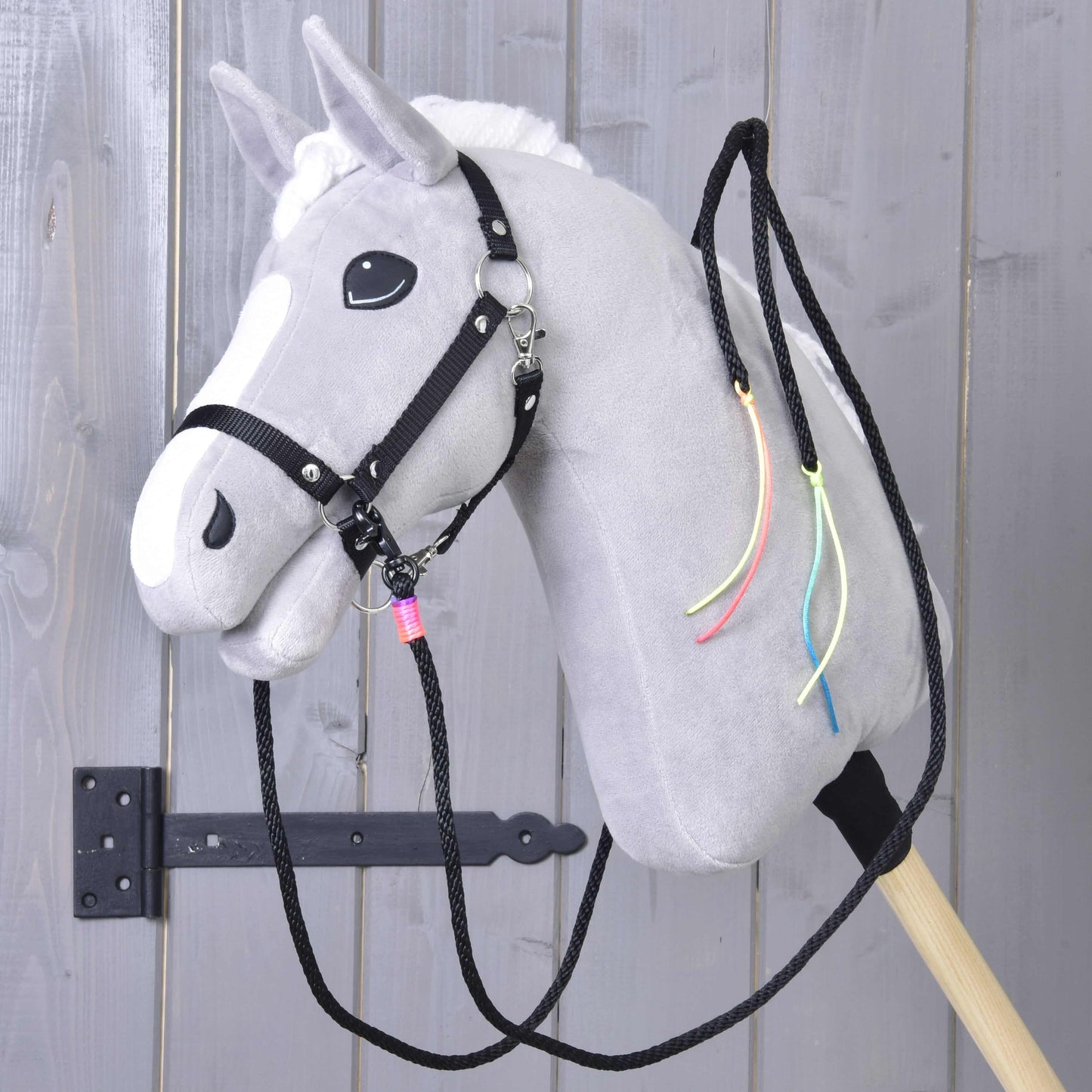 Hobby Horse LarDen "Westernové opraty Texas delené", sivý plyšový koník s bielou hrivou, čiernou uzdečkou, delenými westernovými oprátami a farebnými stuhami na krku, nasadený na drevenej tyči pred sivými drevenými dverami.