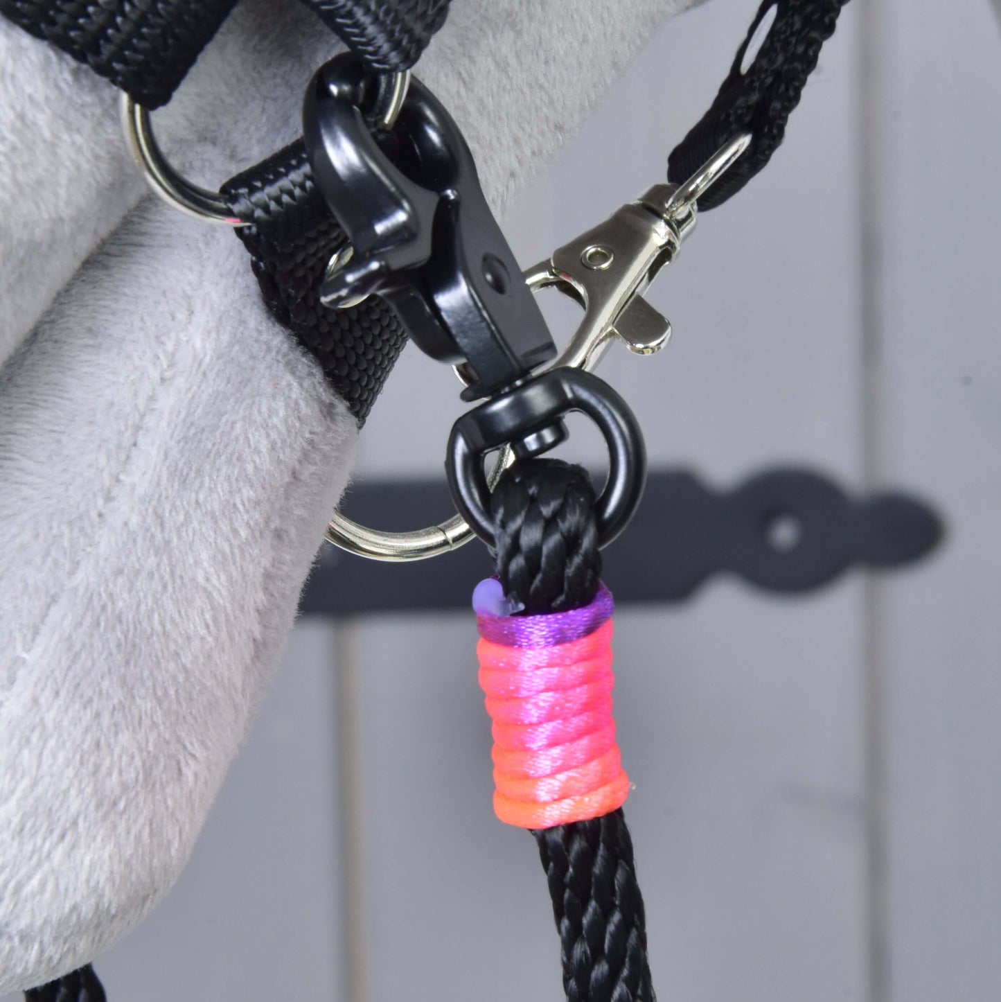Detail Westernových oprát Texas delených od Hobby Horse LarDen s čiernou kožou pripnutou karabínami ku kovovému krúžku na sivom povrchu a s omotaním pri karabíne ružovou, oranžovou a fialovou niťou.
