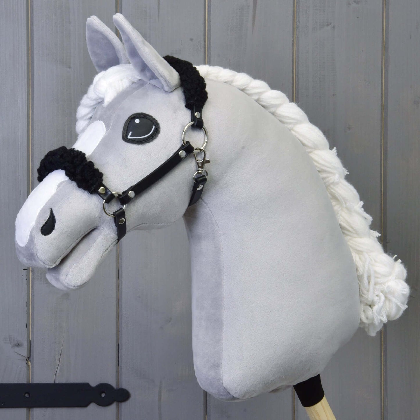 Čierna ohlávka z koženky s čiernym barančekom od Hobby Horse LarDen na plyšovom svetlosivom koni s čiernou koženkovou ohlávkou a veľkými vyšívanými očami, pred sivým dreveným pozadím.