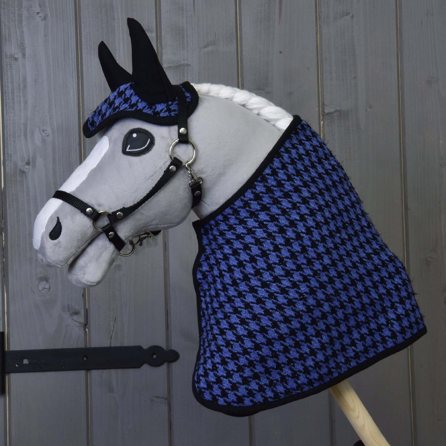 Hobby Horse LarDen s bielou hrivou a čiernou uzdečkou má na sebe Set slávnostná modrá, čabraku a deku so vzorom kohútej stopy, a pózuje pred sivou drevenou stenou.