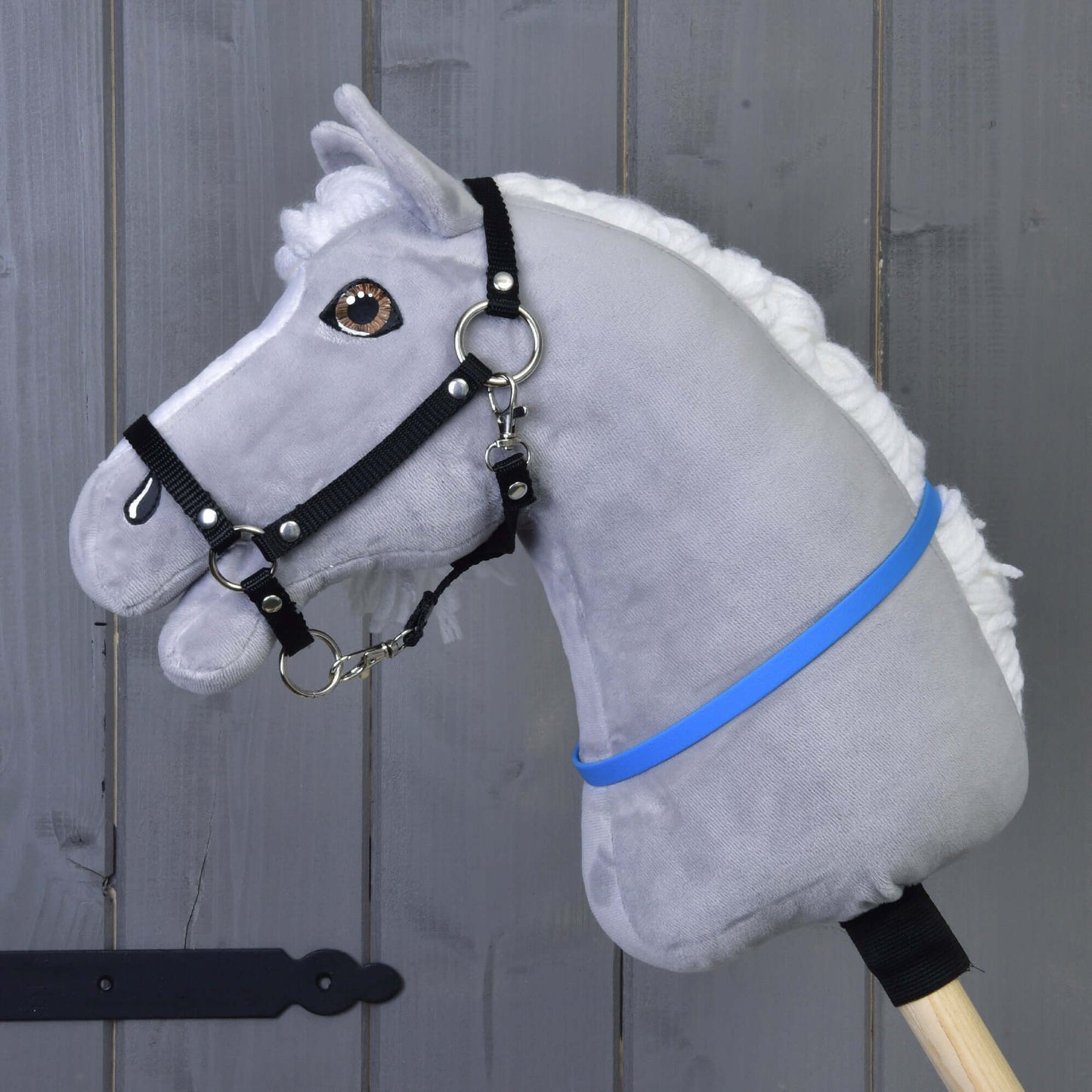 Hobby Horse nákrčník modrý (veľ. S) od Hobby Horse LarDen na sivom plyšovom Hobby Horse s bielou hrivou, čiernou uzdečkou a modrofialovou stuhou na drevenej tyči, pred sivou drevenou stenou.