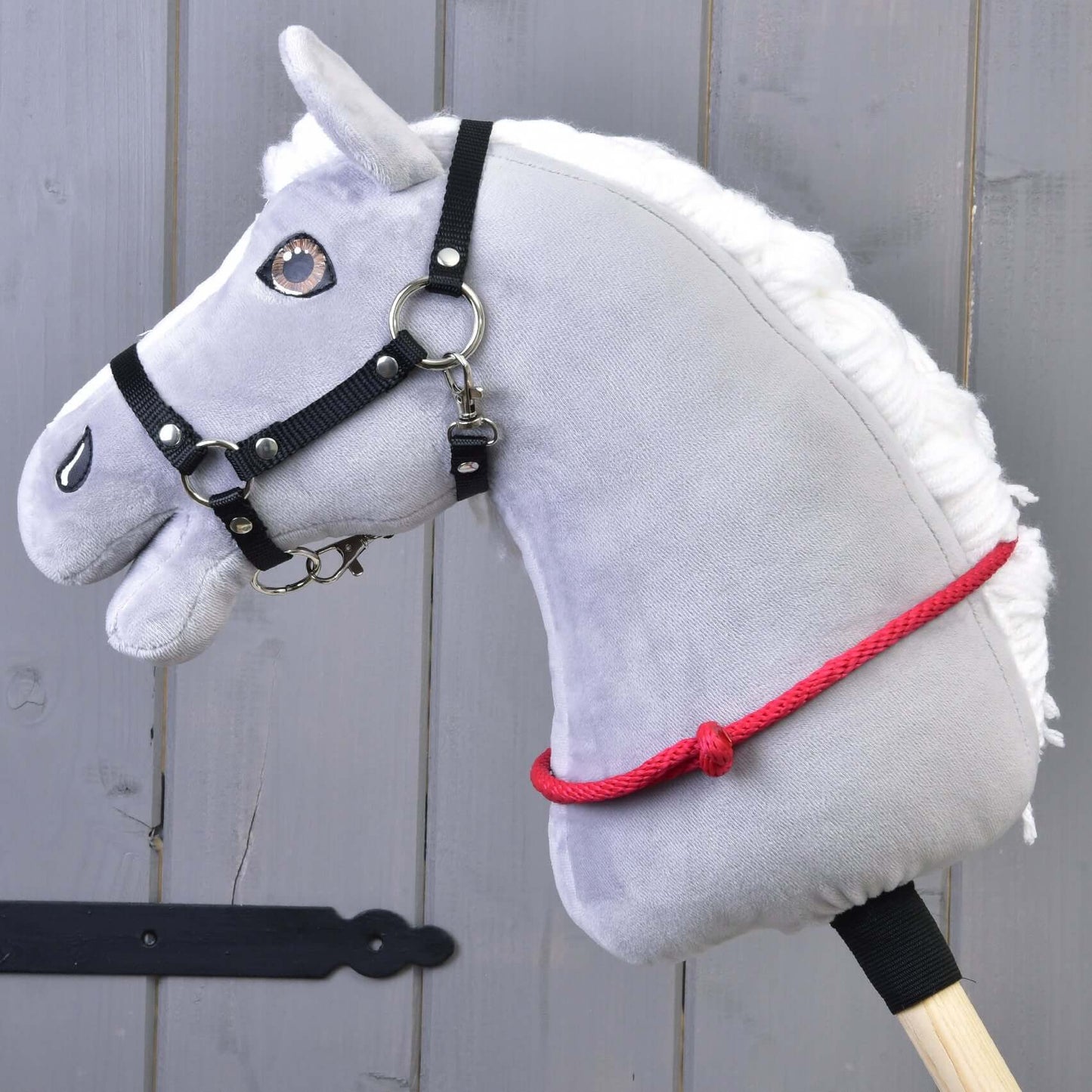 Sivá plyšová hlava Hobby Horse s čiernou uzdečkou, kovovým zubadlom a ručne vyrobeným Nákrčníkom červeným (veľ. S) od Hobby Horse LarDen na drevenej tyči, pred sivou drevenou stenou.