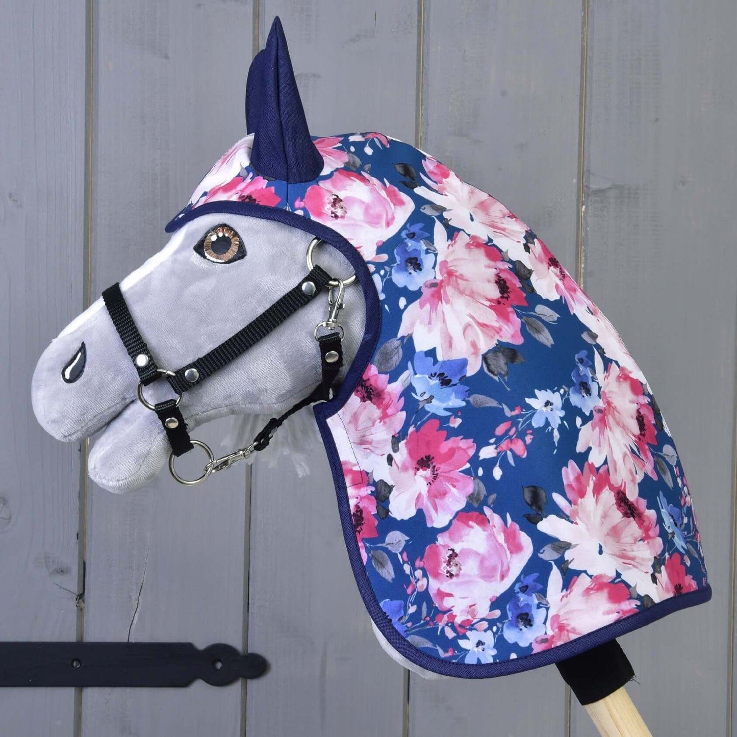 Hobby Horse LarDen „Pláštěnka Letní Pivoňka“ je šedý Hobby Horse s černou uzdečkou, oblečený v modré pláštěnce zdobené růžovými a bílými květy, nasazený na dřevěné tyči před šedou dřevěnou stěnou.