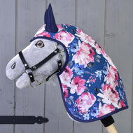Hobby Horse LarDen „Pláštěnka Letní Pivoňka“ je šedý Hobby Horse s černou uzdečkou, oblečený v modré pláštěnce zdobené růžovými a bílými květy, nasazený na dřevěné tyči před šedou dřevěnou stěnou.