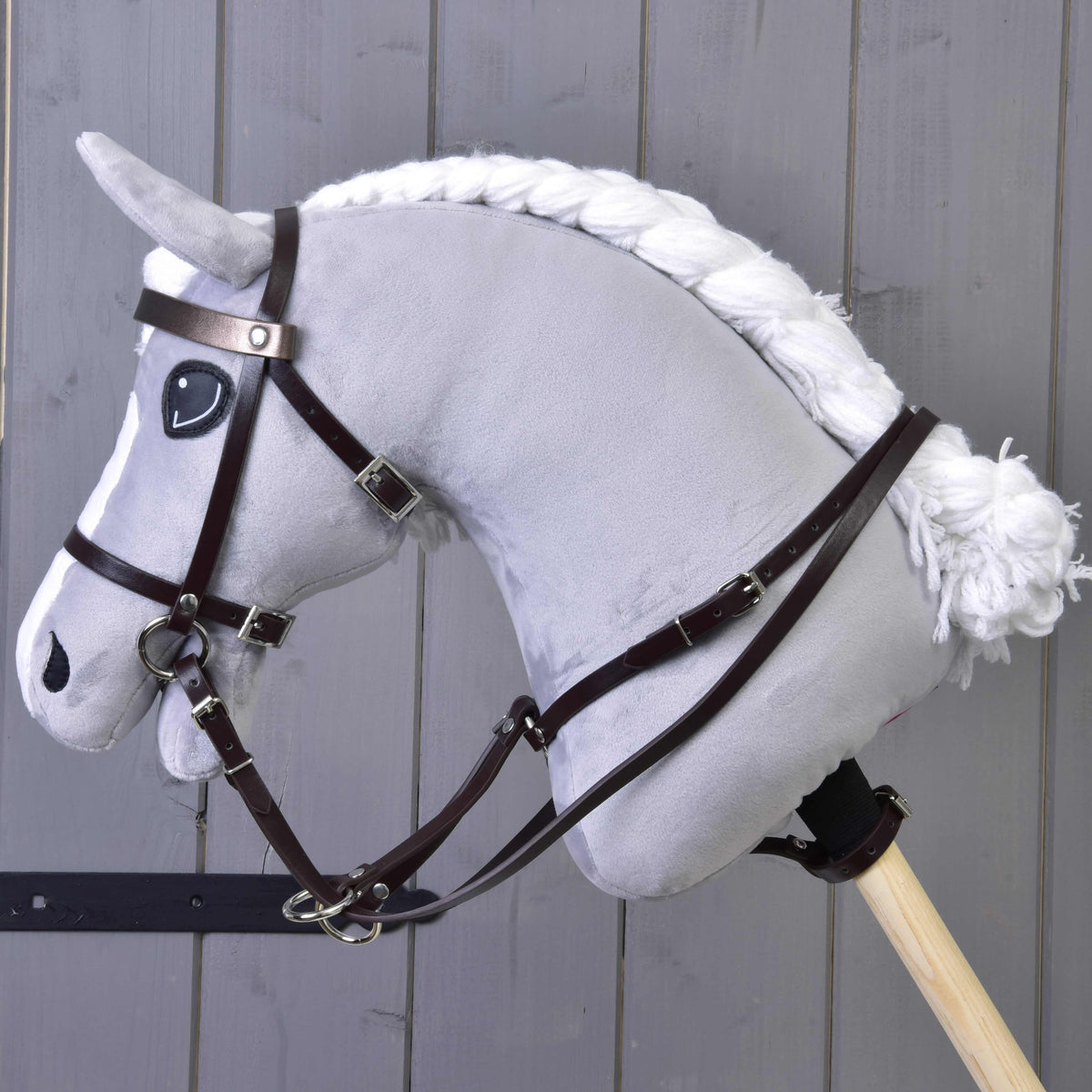 Hobby Horse LarDen Martingal hnedý, sivý plyšový Hobby Horse s bielou priadzovou hrivou a chvostom, čiernou uzdečkou a koženkovým martingalom na drevenej tyči, je zobrazený pred pozadím zo sivých drevených dosiek.