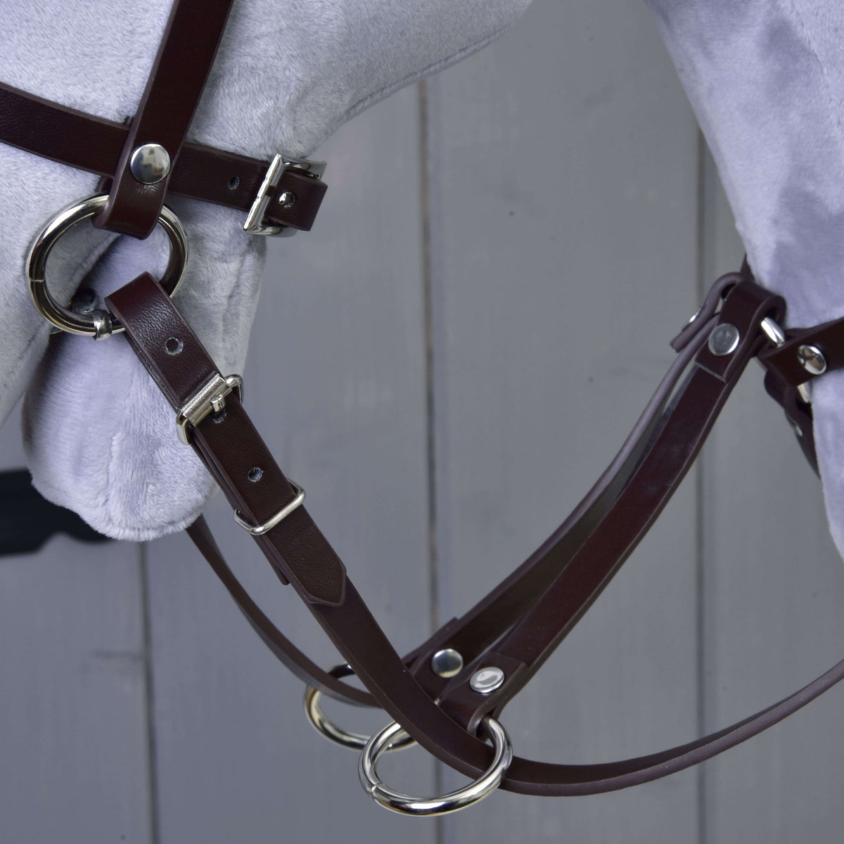 Detail Martingal hnedý od Hobby Horse LarDen s hnedou koženou uzdečkou s kovovými prackami a krúžkami na svetlosivej hlave modelu koňa na pozadí sivej drevenej steny.