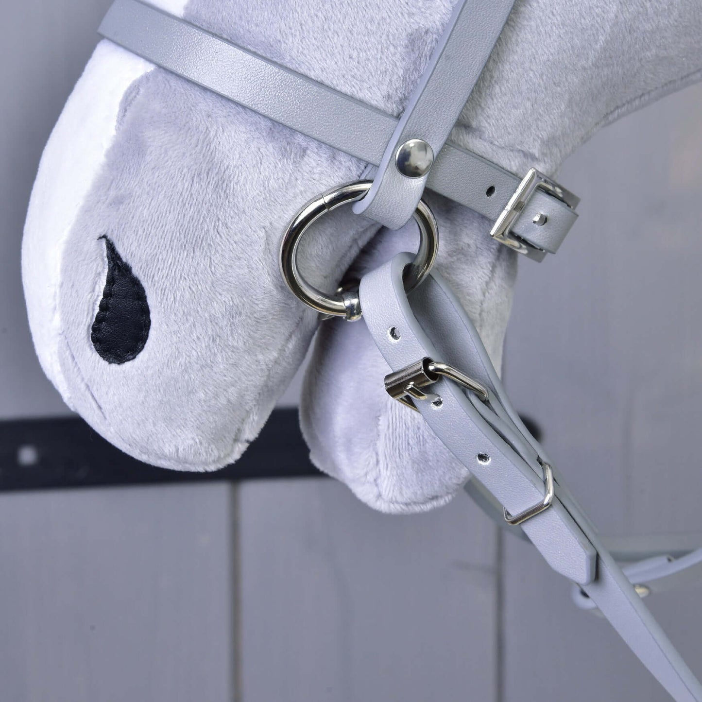 Detailní záběr na šedého plyšového Hobby Horse od Hobby Horse LarDen se Šedým koženkovým setem - uzdečka, otěže a martingale, s kovovými kroužky a přezkami; černé oči jsou vyšité, v pozadí rozostřené dřevěné obložení.